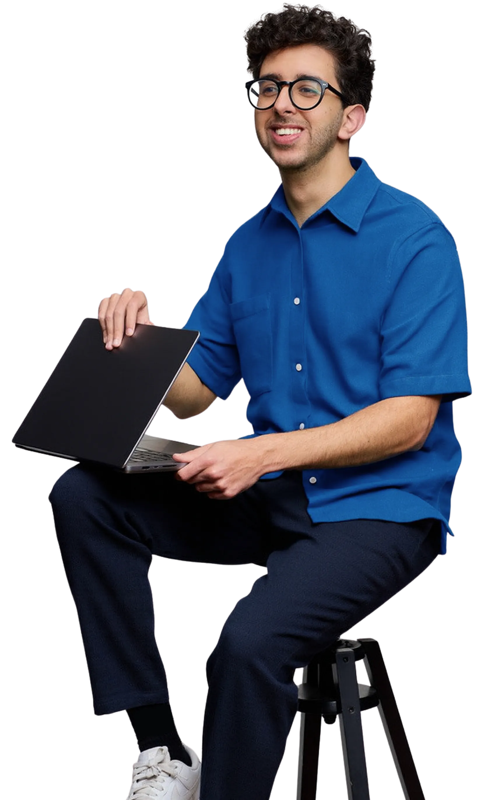 Photo of Oly smiling, sitting on a stool with a laptop wearing a blue shirt and glasses.