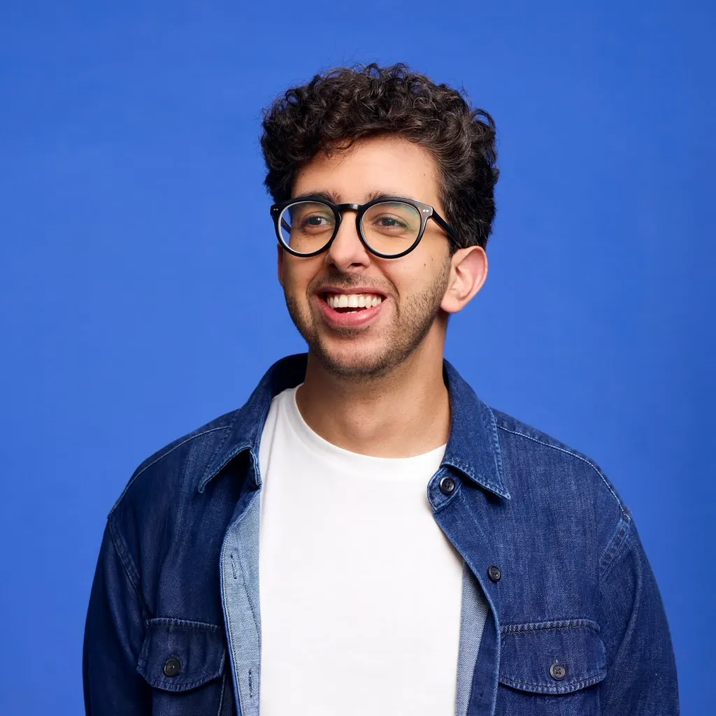 Oly smiling in front of a bright blue background, wearing glasses, a white shirt, and a denim jacket.